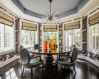 Bold dining space with large bay window, blue painted ceiling, black dining table, dark wooden flooring, patterned wallpaper, windows dressed with blinds, sculptural light fixture