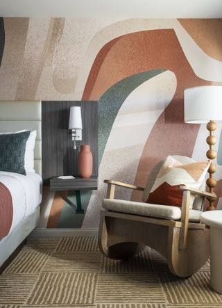 A whimsically furnished hotel bedroom features wide pastel-shaded wall murals, a curvilinear wooden, playful armchair, a standing lamp in a similar aesthetic, and a carpeted floor in sandy tones.