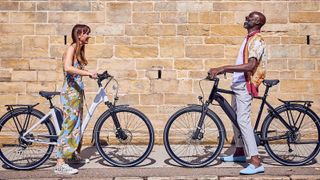 Two cyclists standing over their electric bikes face each other while laughing