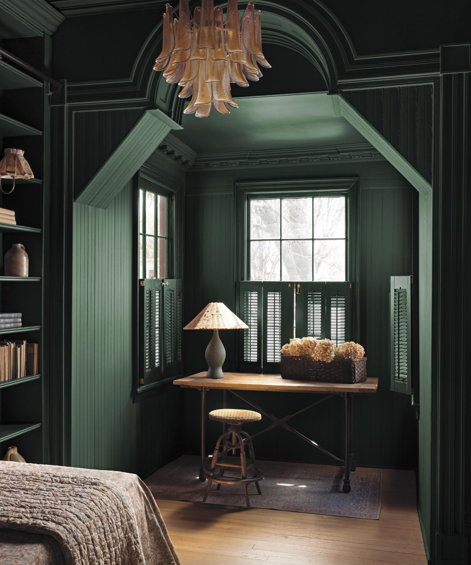 A cosy bedroom painted in Webster Green by Benjamin Moore with an arched alcove and a desk by the window, wooden floors, built-in shelves filled with books, and a glass chandelier hanging above.