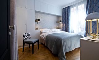 Interior view of a bedroom at the Seine side apartment featuring wood flooring, a bed with white pillows and white and grey linen, a white wall-mounted unit with a wardrobe, wall lights, a window with blue patterned curtains and a chair with a black and white geometric patterned backrest