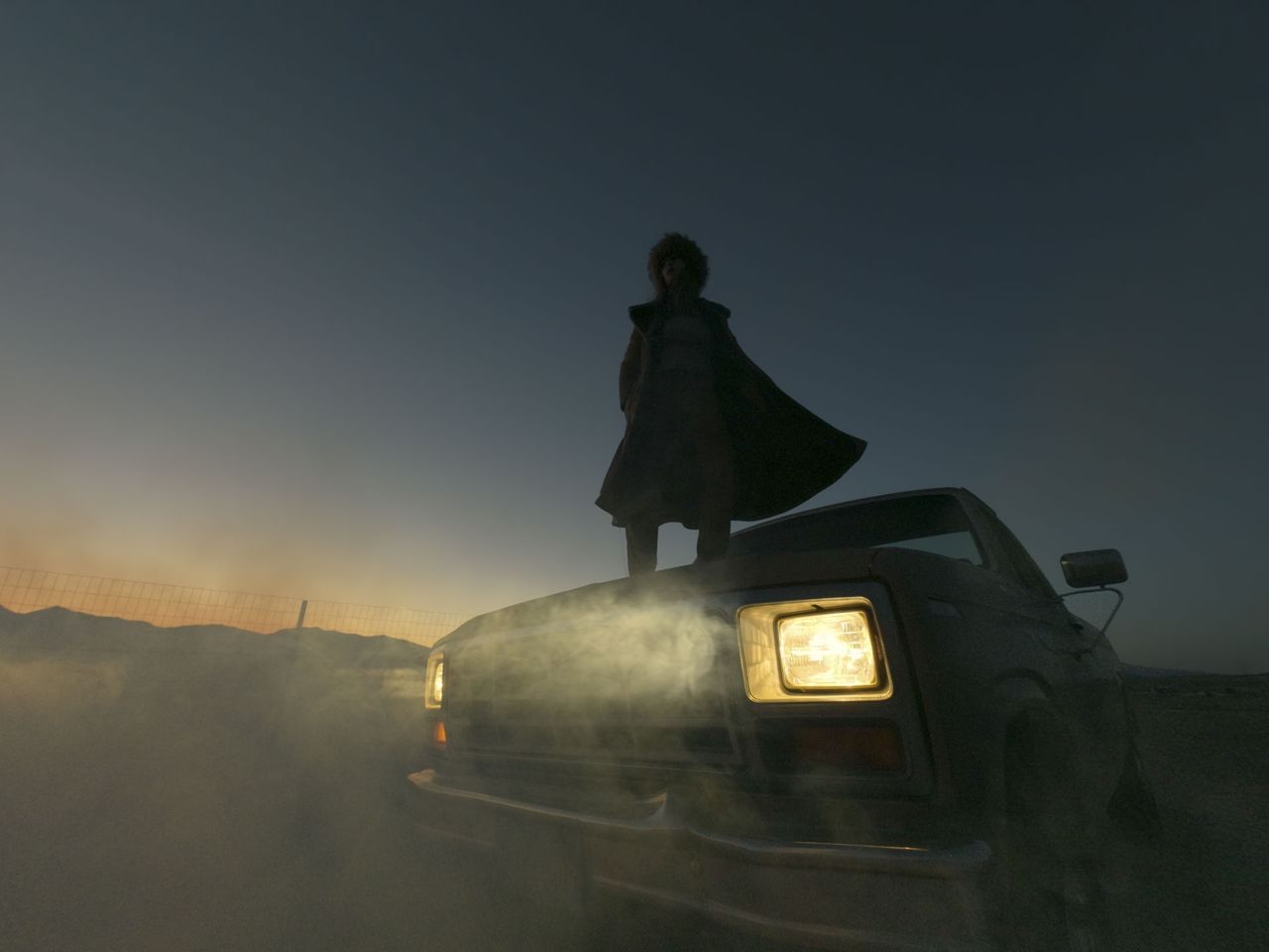 Female model standing on a car bonnet at dusk, the car's headlamps are on