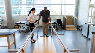 Female physical therapist helps a senior man walk following a stroke. The man is using parallel bars in a rehab center.