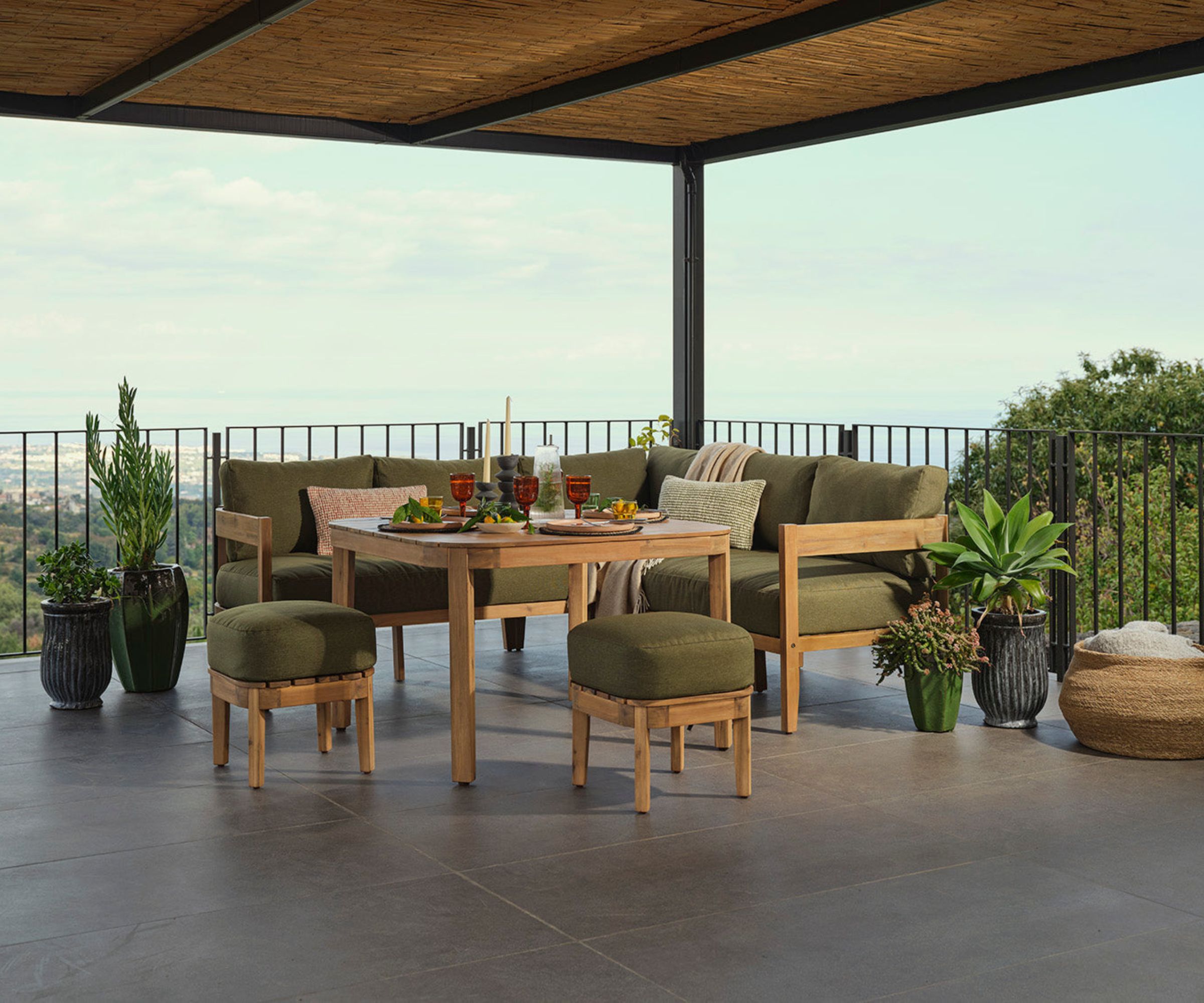 Wooden corner dining set with olive cushions on covered terrace, table set for dinner, hillside sea view.