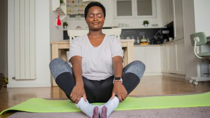 woman wearing a white tshirt and leggings sitting on the floor on a green yoga mat in butterfly pose, facing the camera with eyes closed. there's a kitchen behind her. 