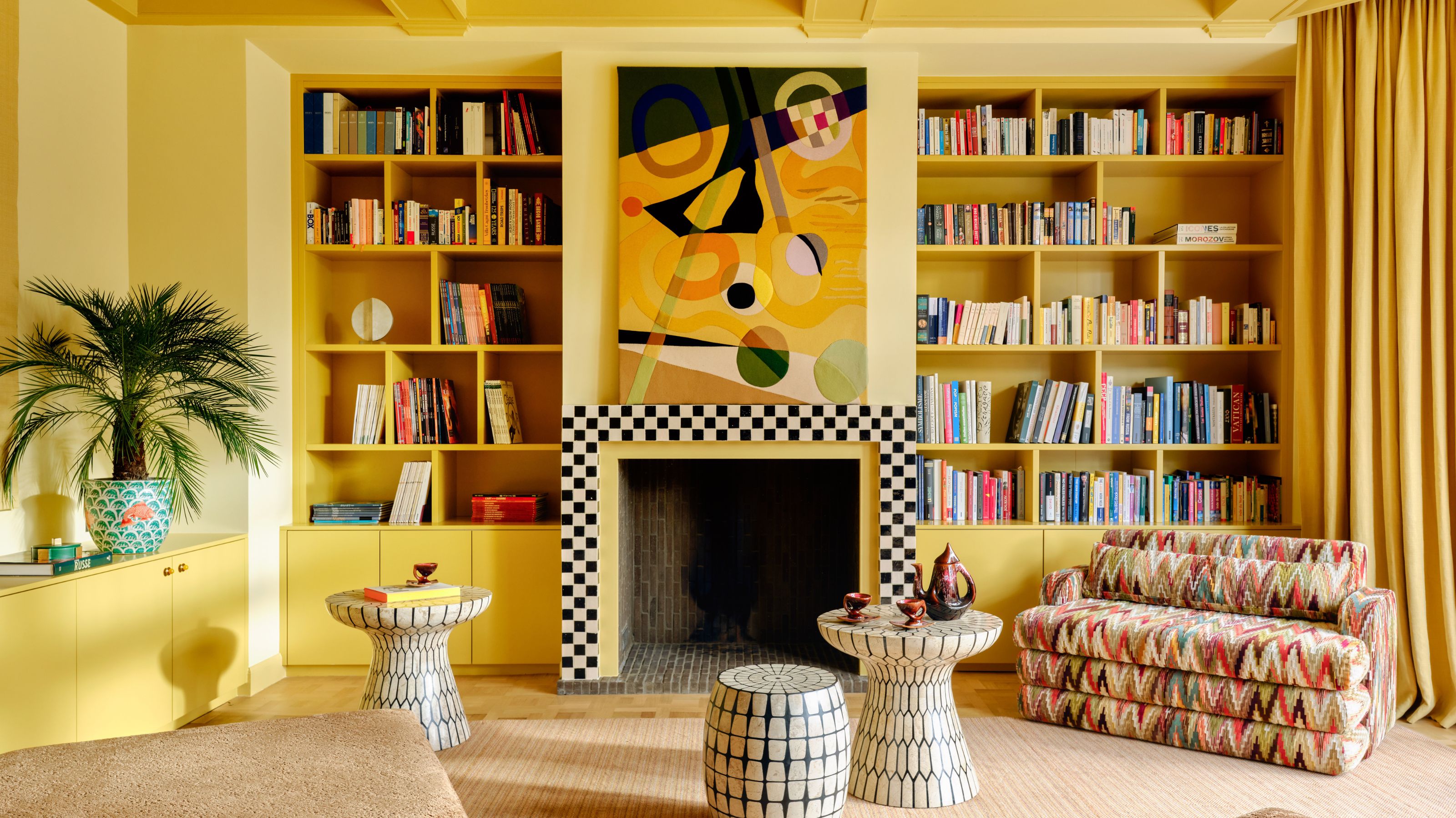 a bright yellow painted living room with a checkerboard tiled fire surround, bookcases, and eclectic furniture and art