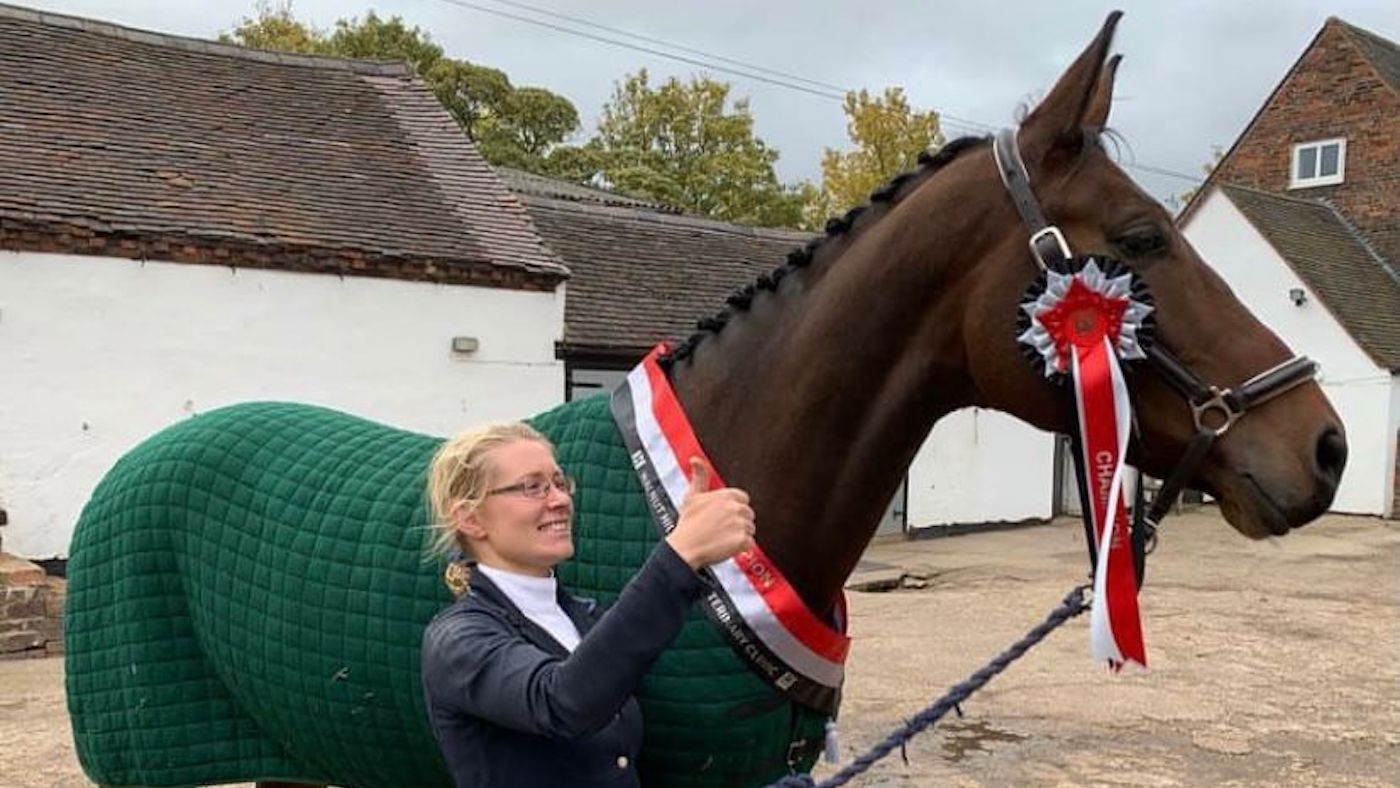 Claire Holliday giving a thumbs up sign, as she holds her horse, Frankie, who is wearing a green rug and a red rosette and sash