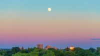moon shines above a city scape. the sky is colored hues of blue, orange, yellow and violet.