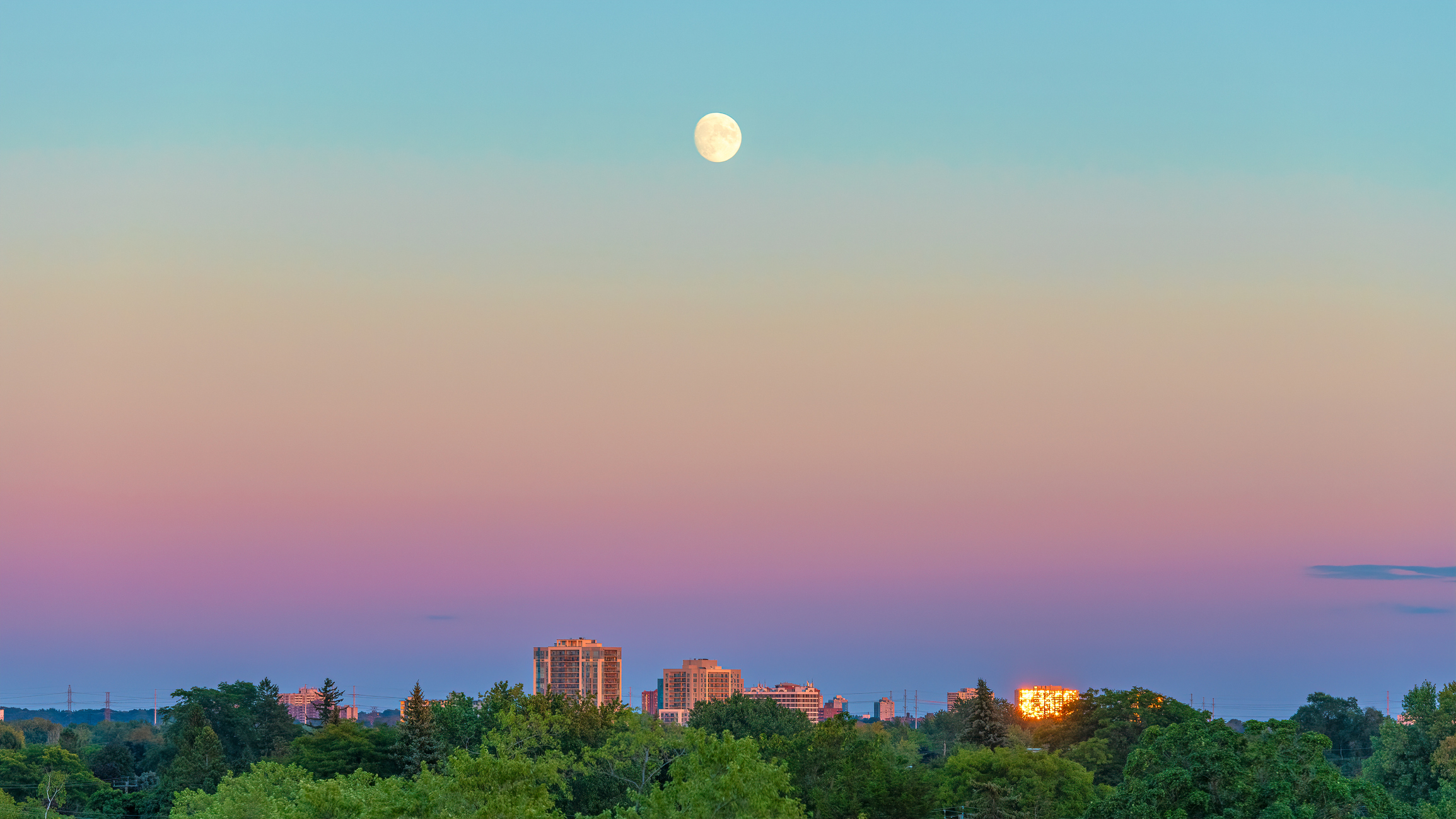 moon shines above a city scape. the sky is colored hues of blue, orange, yellow and violet.
