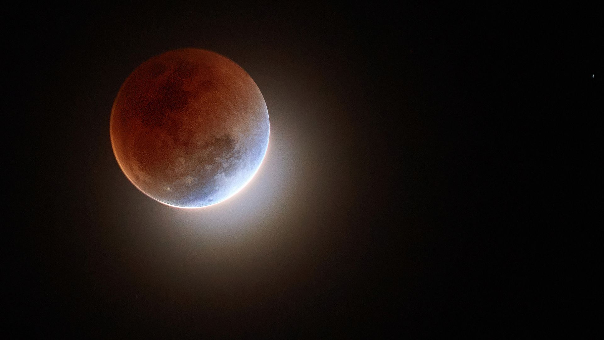 Image of a red-tinted moon against a clear black sky with a small ring of light peeking out from the bottom right of the moon. 