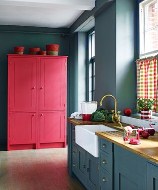 Dark blue painted kitchen with bright red kitchen freestanding wooden cabinet, light wooden flooring, sink with brass tap, multi-colored gingham half curtains