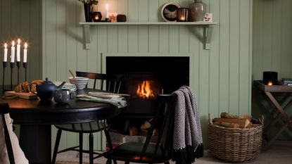 Cozy sage green panelled living room with a lit fire in the fireplace, a circular black dining table with lit candles in a candelabra, and a large wicker basket filled with logs.