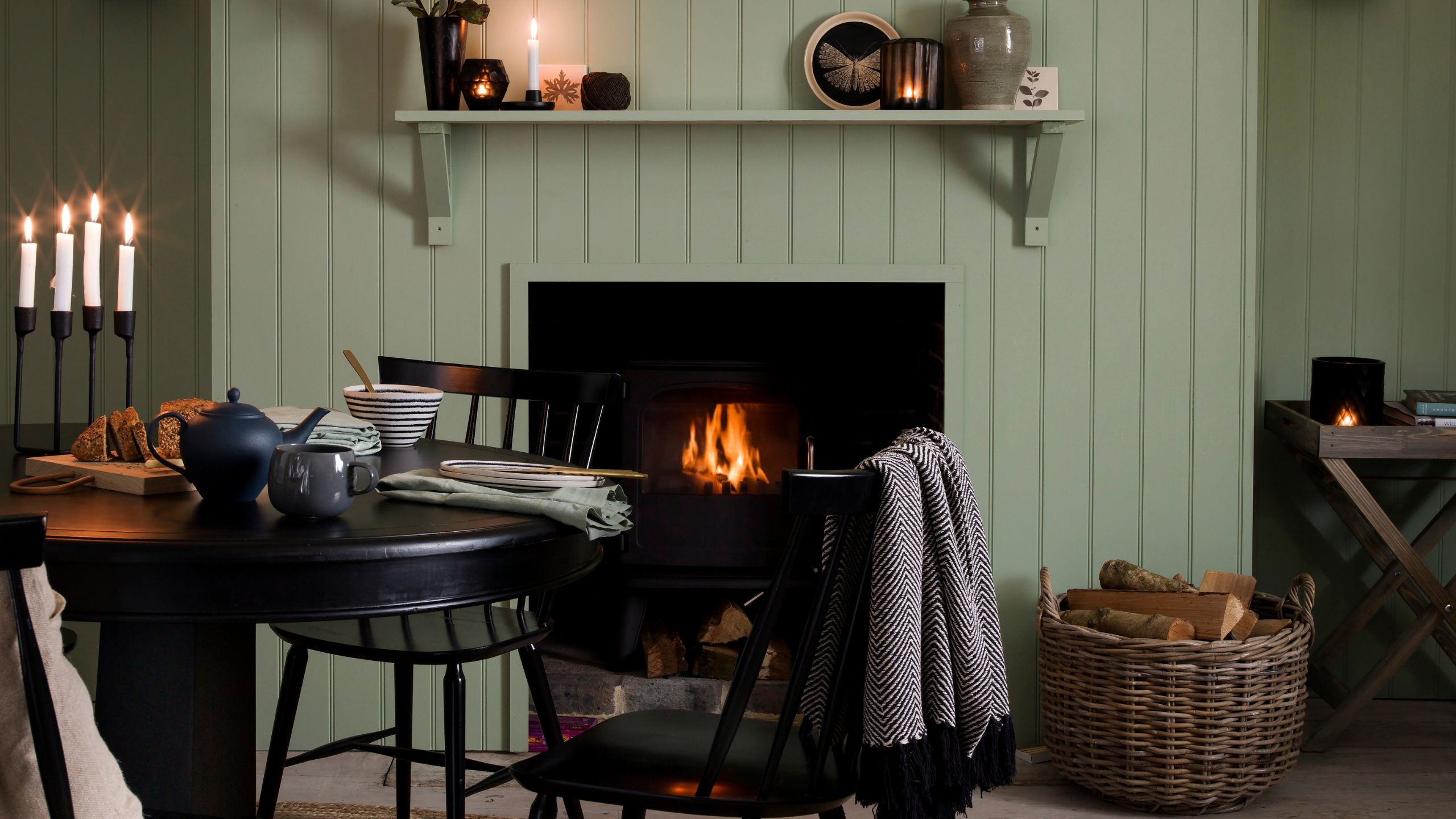 Cozy sage green panelled living room with a lit fire in the fireplace, a circular black dining table with lit candles in a candelabra, and a large wicker basket filled with logs.