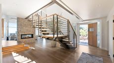 A large wooden entryway flooding with natural light from an open wooden door, with a central spiralling staircase, an exposed wood accent wall with a wood burner, and steps up to a comfortable living area seen in the background of the left hand corner.