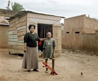 An African woman standing next to a boy with his foot on a skateboard and a shack behind them.
