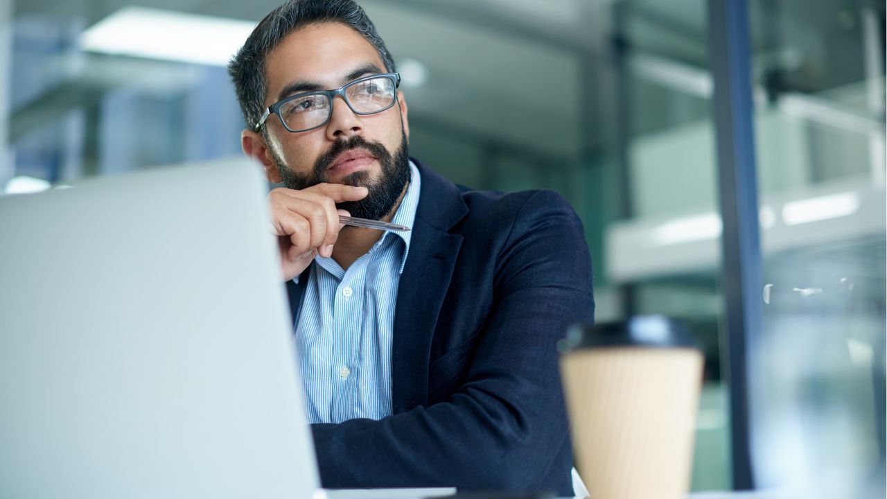 An office worker sits at his desk in front of his computer and appears to be thinking.