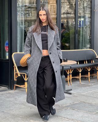 Salome wears long gray wool duster coat, low-rise black relaxed trousers, cropped tee, and Sambas.