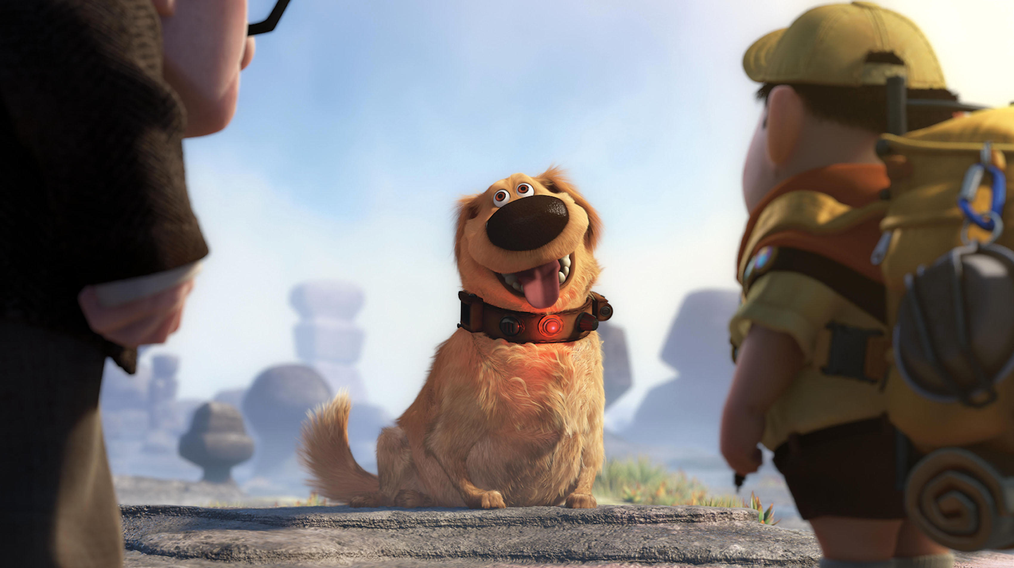 An animated golden dog with a big nose, floppy ears and a talking collar sits happily with its tongue out, looking up at two characters standing on either side — an elderly man on the left and a young boy scout with a backpack on the right — in a rocky, soft-focus landscape under a bright sky.