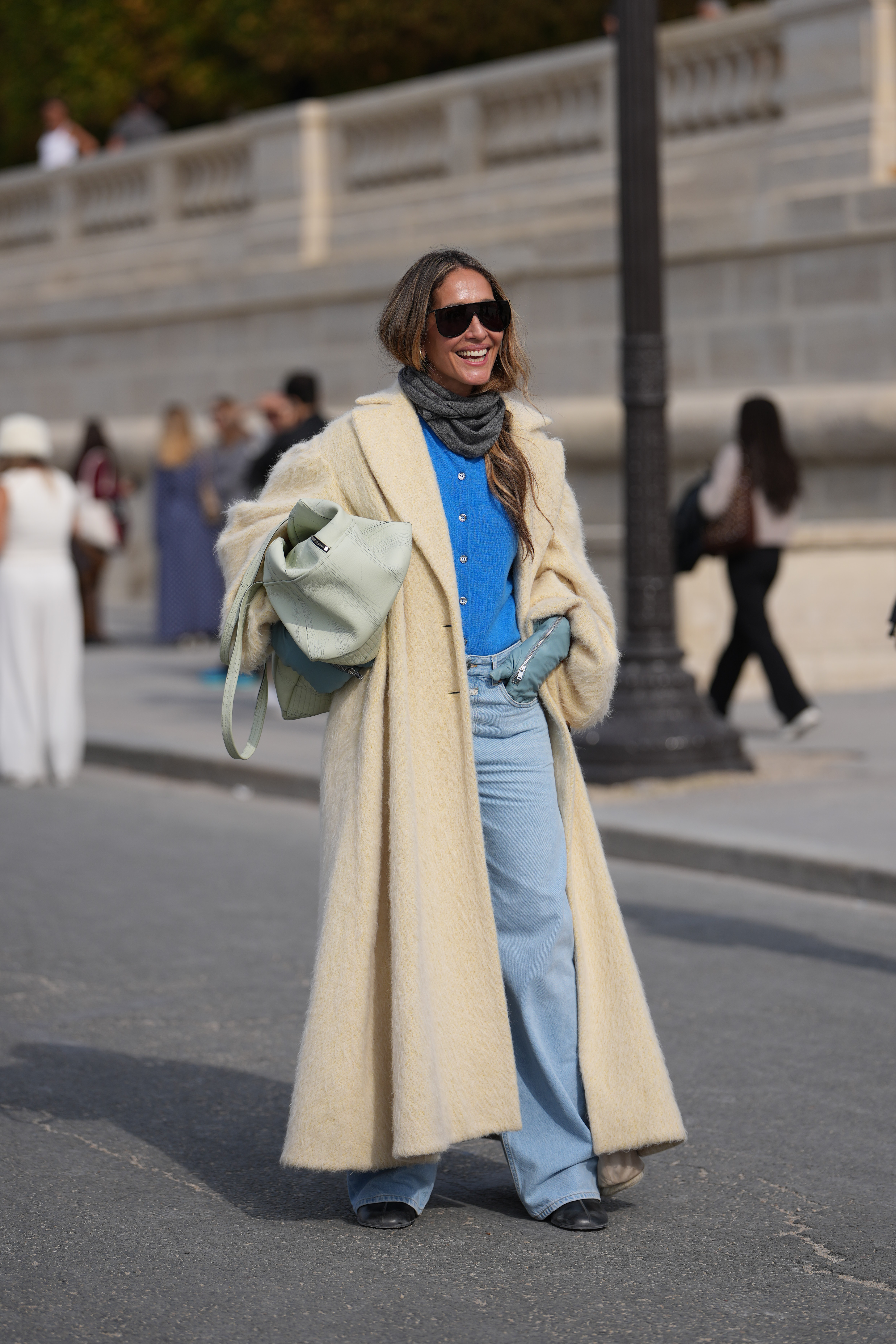 PARIS, FRANCE - OCTOBER 01: A guest wears long wavy light brown hair with a center part, oversized black rectangular sunglasses, small gold earrings, a pale mint green leather puffy shoulder bag, a grey knit scarf, a bright cobalt blue button-up knit cardigan, a long cream fuzzy coat with a light satin lining, pale blue leather gloves, light wash high-rise wide-leg denim jeans, black leather boots shoes with a low heel, outside Dior, during Paris Fashion Week - Womenswear Spring Summer 2026, on October 01, 2025 in Paris, France (Photo by Edward Berthelot/Getty Images)