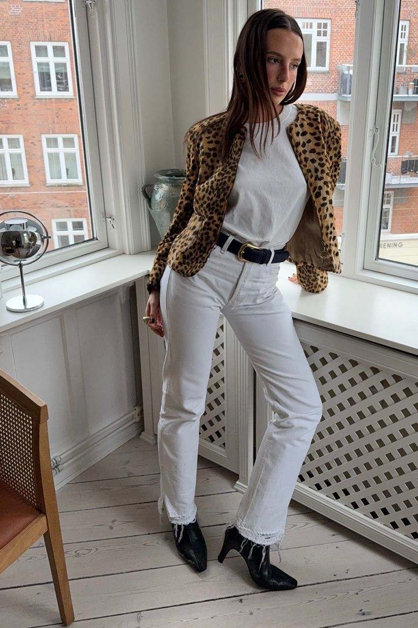 A woman standing inside an apartment by the windows wearing a collarless cheetah print pony-hair jacket styled with a white t-shirt, a black belt, white straight-leg jeans, and black ankle boots.