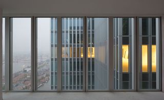 View from a high rise window at De Rotterdam , white walls and floor, tall windows, view of adjacent wings of the building with some lights on, surrounding landscape to the bottom left, buildings, water, roads, cars, trees , grey misty sky