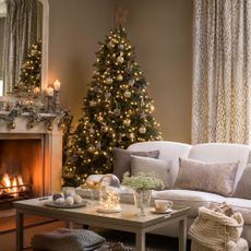 Decorated Christmas tree in a country style brown living room, white sofa, cushions, fireplace, lit fire, decorated mantelpiece, large mirror, coffee table
