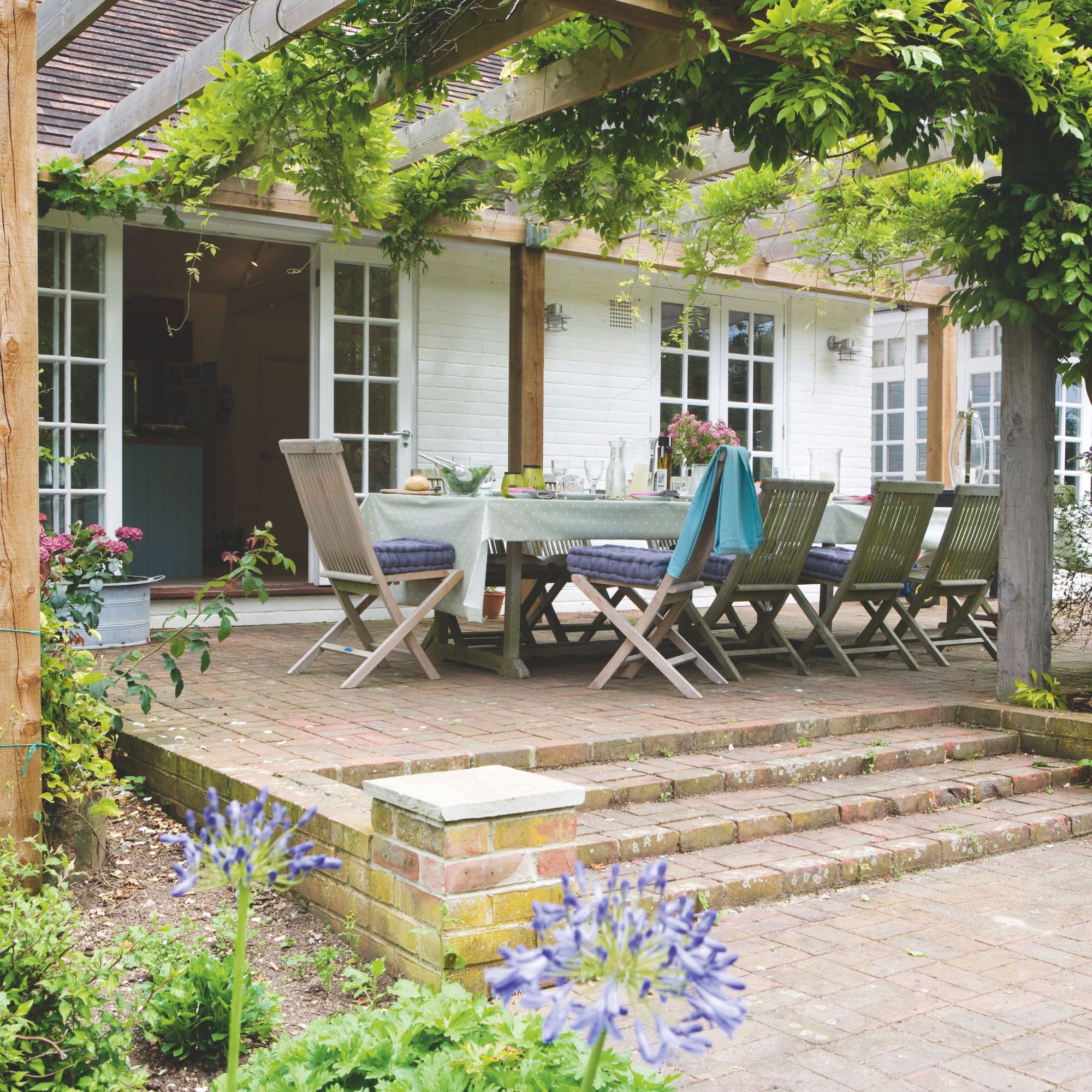 Outdoor seating area beneath wooden pergola with climbing plants and wall lights on house
