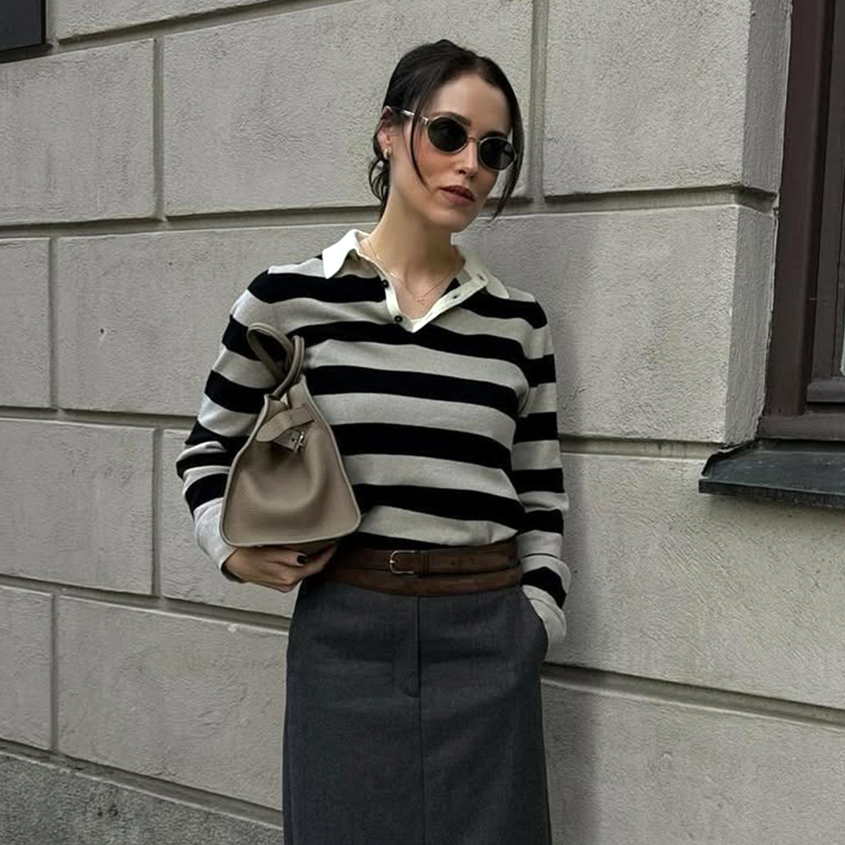 Influencer Anna wears a striped polo sweater and a grey skirt with a brown belt and black loafers and light colored leather bag. 