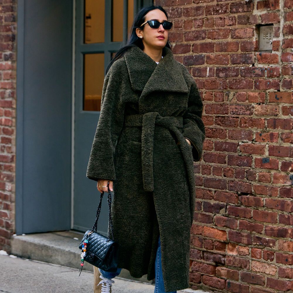 Woman wears green faux fur coat while carrying a black quilted handbag and tan sneakers. 
