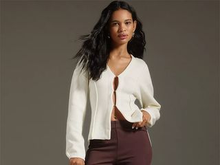 Model wears white cardigan and brown cropped pants while holding a black clutch.