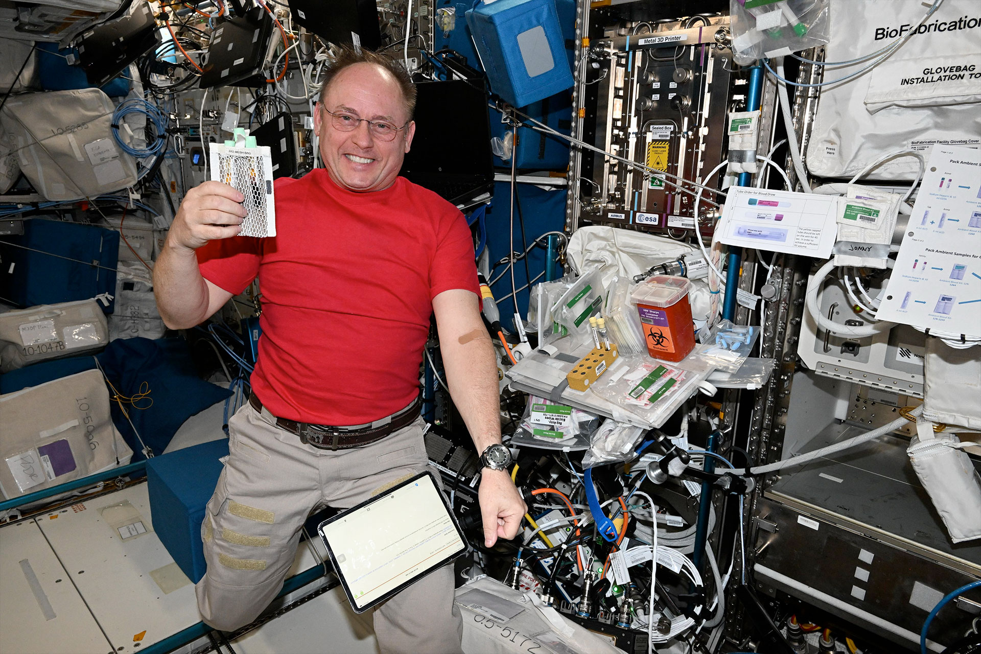 A man in a bright red short-sleeve shirt and khaki pants holds up a medical sample bag while floating inside a space station module