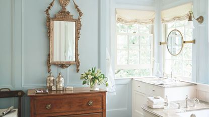 Traditional bathroom with pale blue painted walls, mahogany sideboard with an ornate gold mirror above it, marble bathtub and vanity unit with sink, and two windows with cream roman blinds
