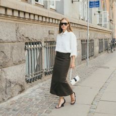 Woman wears black pencil skirt, white button-down top, black wedges, and white small handbag while posing for the camera. 