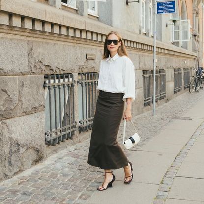 Woman wears black pencil skirt, white button-down top, black wedges, and white small handbag while posing for the camera. 