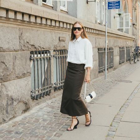 Woman wears black pencil skirt, white button-down top, black wedges, and white small handbag while posing for the camera. 