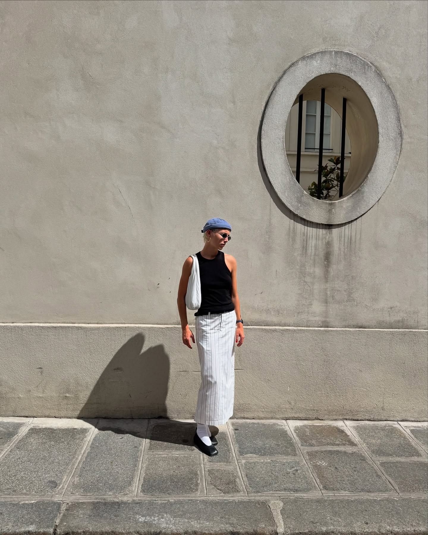 Emma Chamberlain stands outfit wearing a white long pencil skirt with a black tank top and a blue hat.