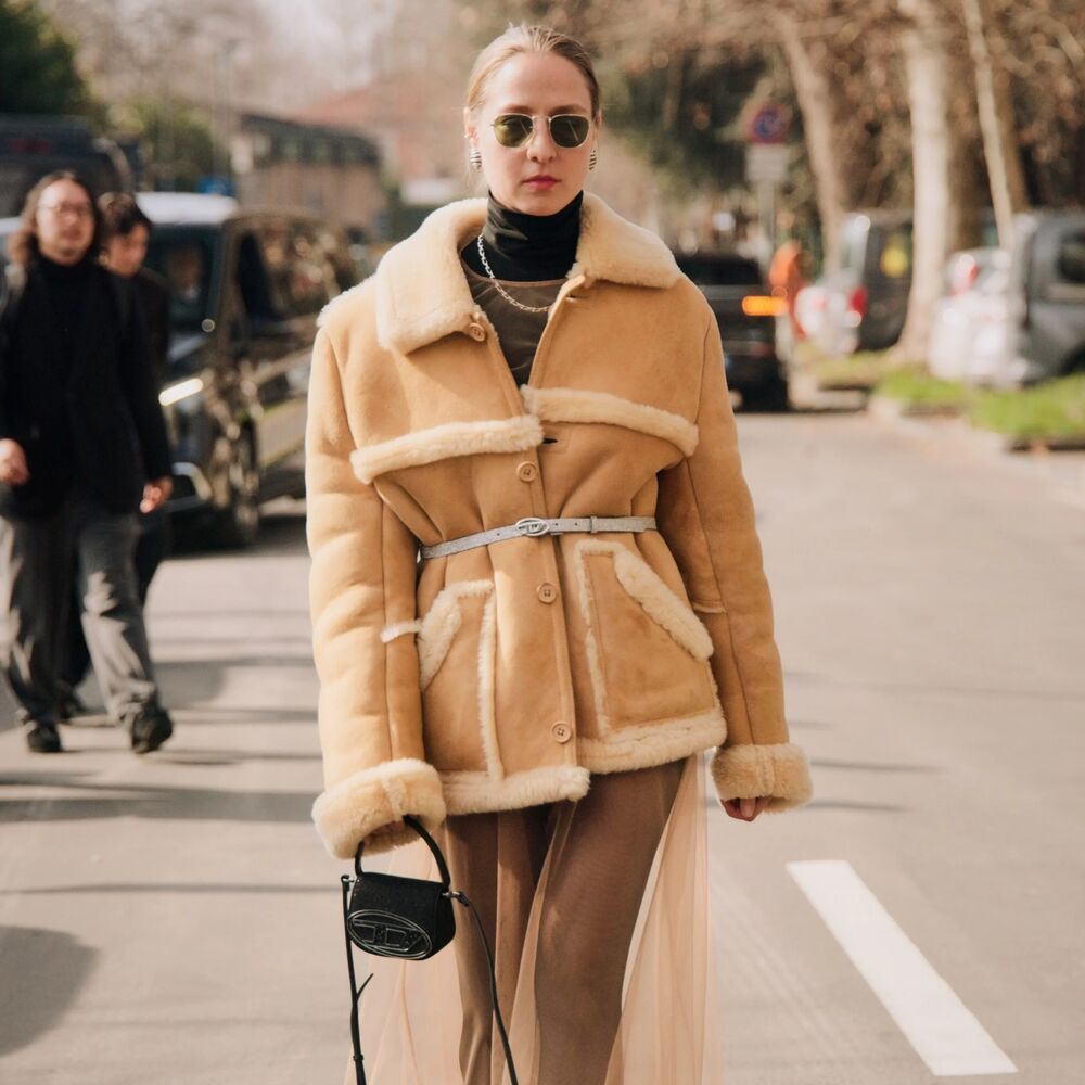 Woman wears fur trimmed suede jacket with sheer maxi skirt and small black handabag