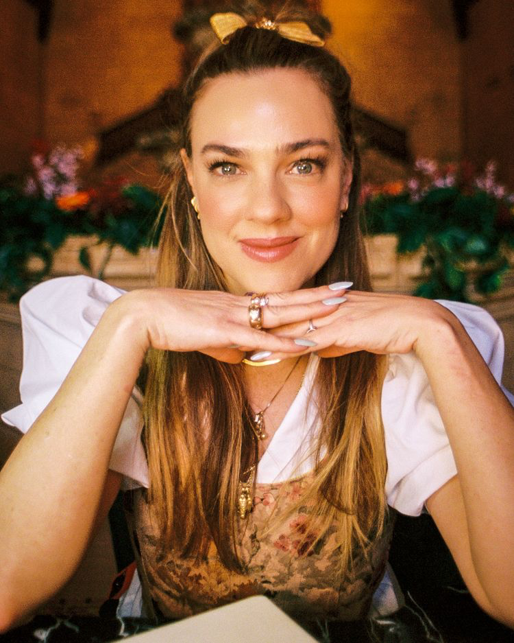 a headshot of a white woman with bronde hair posing with her hands under her chin