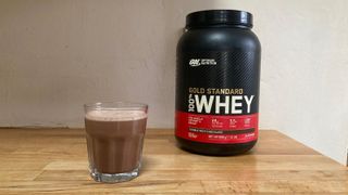 Tub of Optimum Nutrition Gold Standard 100% Whey double chocolate flavour next to small glass of a brown protein shake