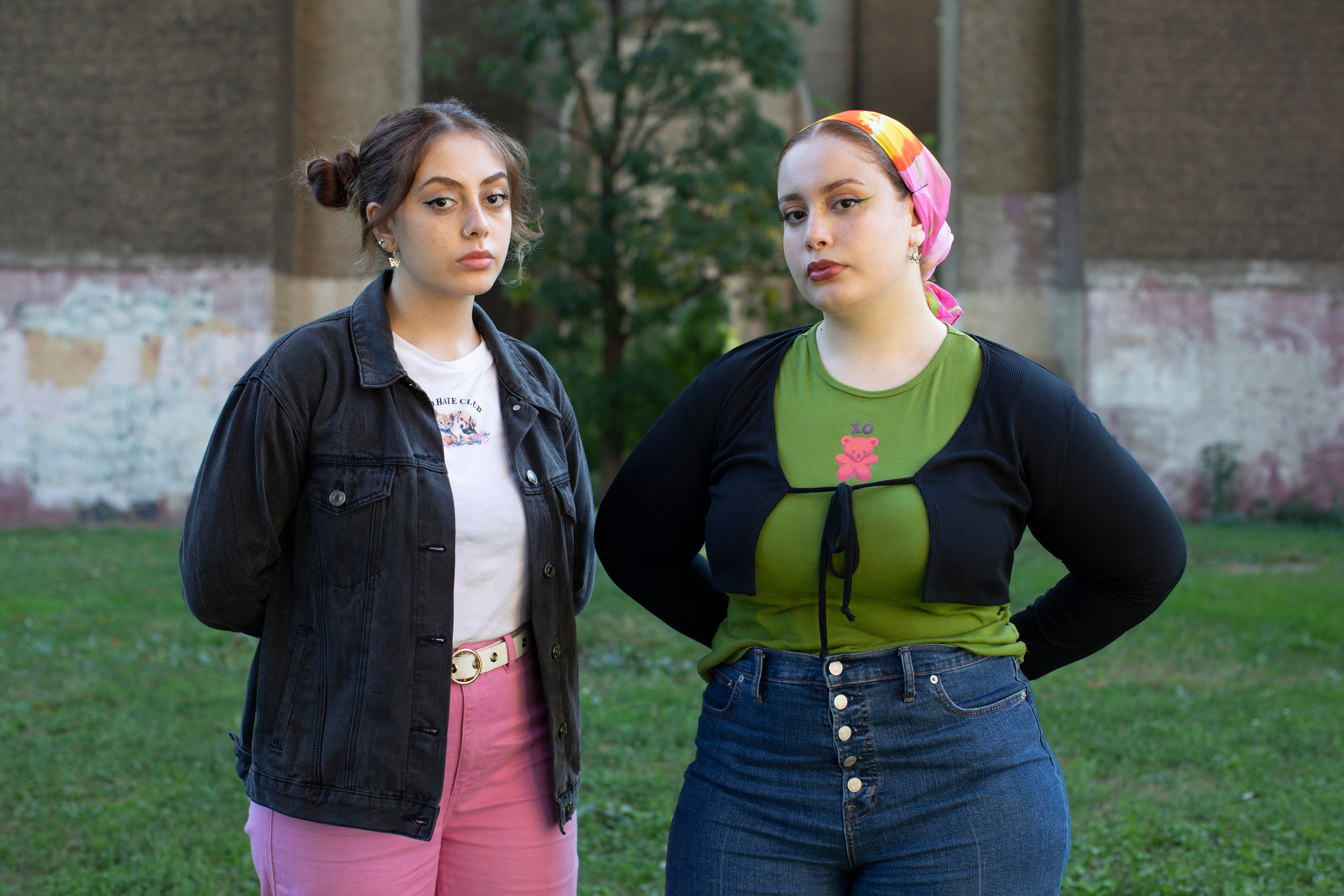 Two young women stand side by side in front of a weathered stone wall: one in a black denim jacket and pink jeans, the other in a green crop top, black cardigan and colourful headscarf, both looking directly at the camera.