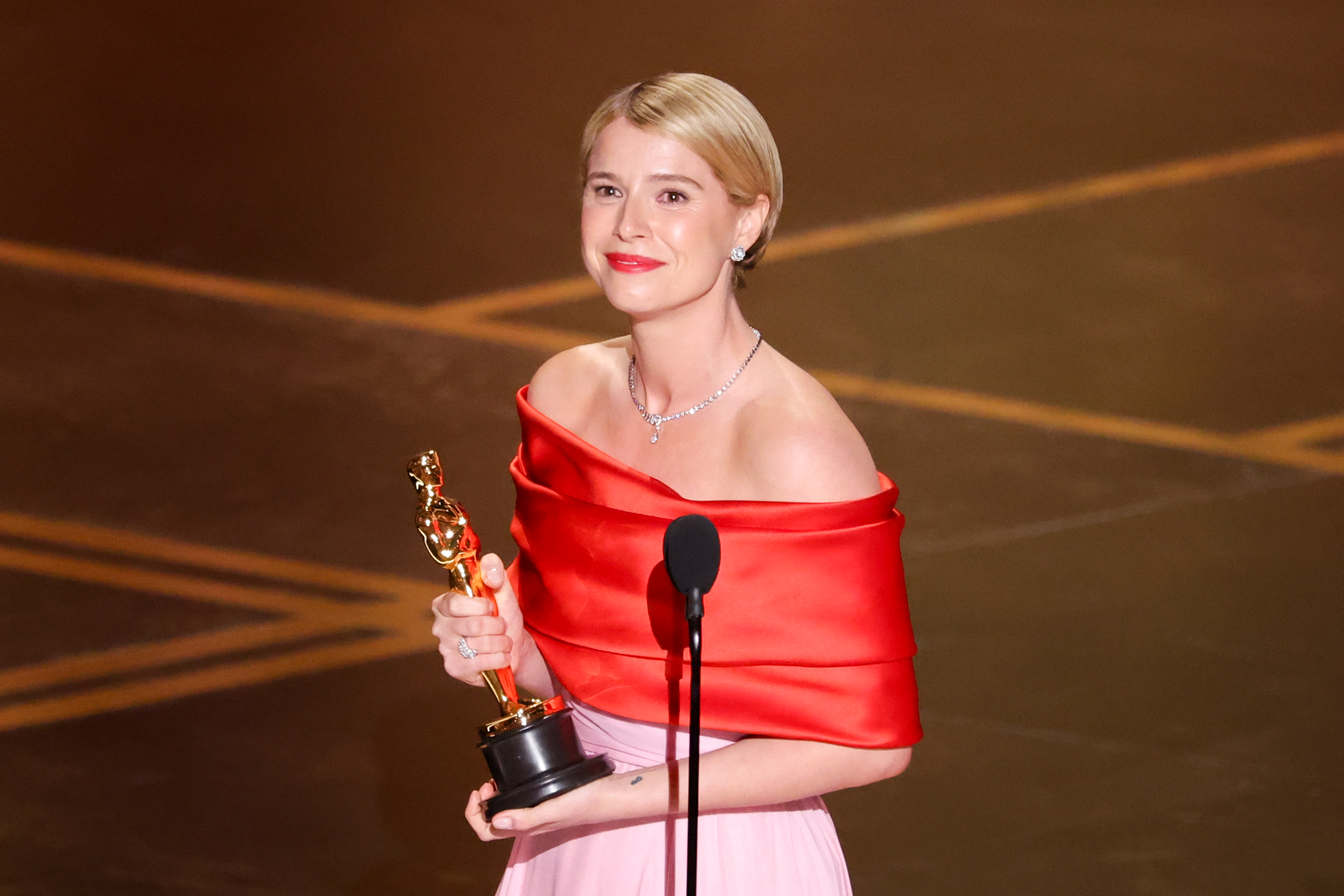 Jessie Buckley wears a red and pink gown and looks emotional as she accepts the award for best actress at the 98th Annual Oscars held at Dolby Theatre on March 15, 2026 in Hollywood, California