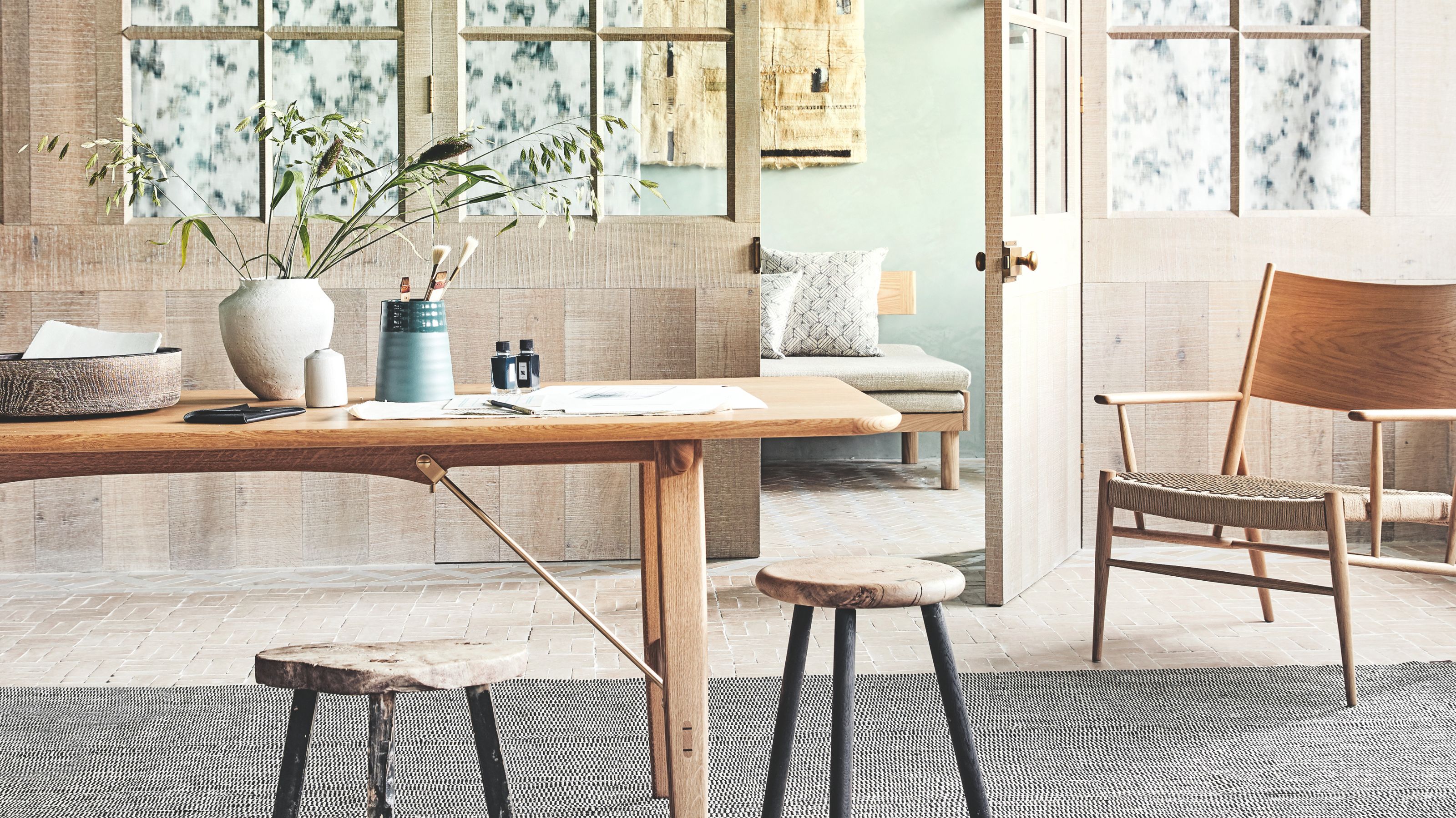 Dining room with wooden table, chair and stools on a grey rug and wooden clad and green walls.