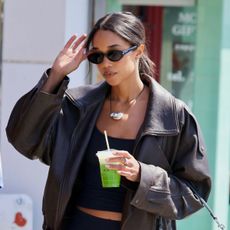 Laura Harrier walking in Los Angeles with a matcha wearing a brown leather jacket, cropped black camisole, cropped black trousers, and black Repetto ballet flats.