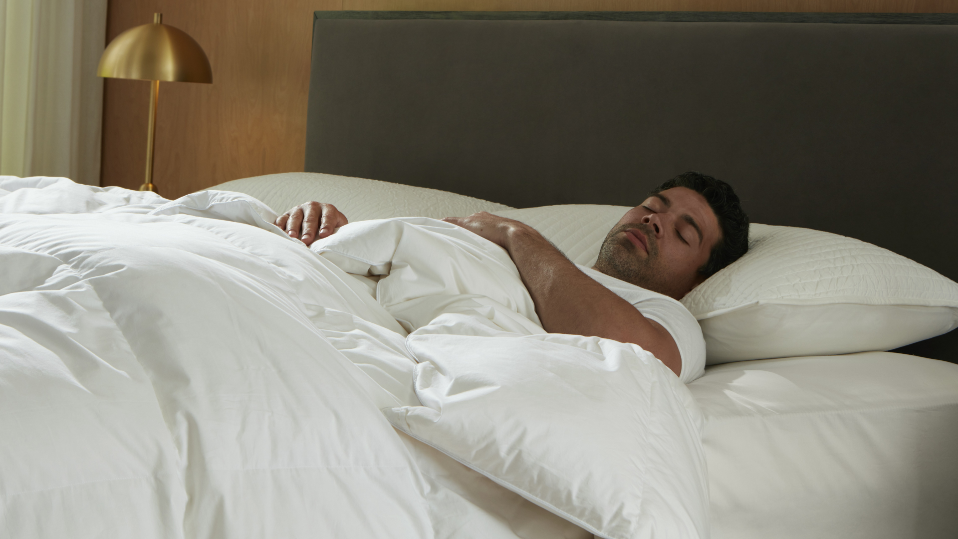 Man sleeping in bed wrapped in the new Saatva Down Bedding collection comforter, pillow and topper