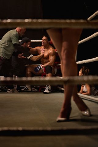 A boxer (Dwayne "The Rock" Johnson) sits in the corner of a ring as two men tend to him. Seen from a distance, with a pair of women's legs and the ropes in the forefront, in 'The Smashing Machine.'