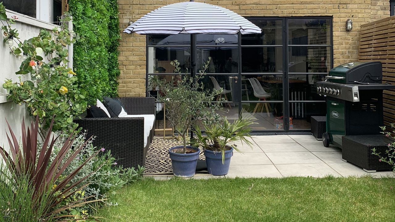 An image of a backyard with lawn laid with blue and white striped parasol umbrella, potted plants, barbecue grill in corner