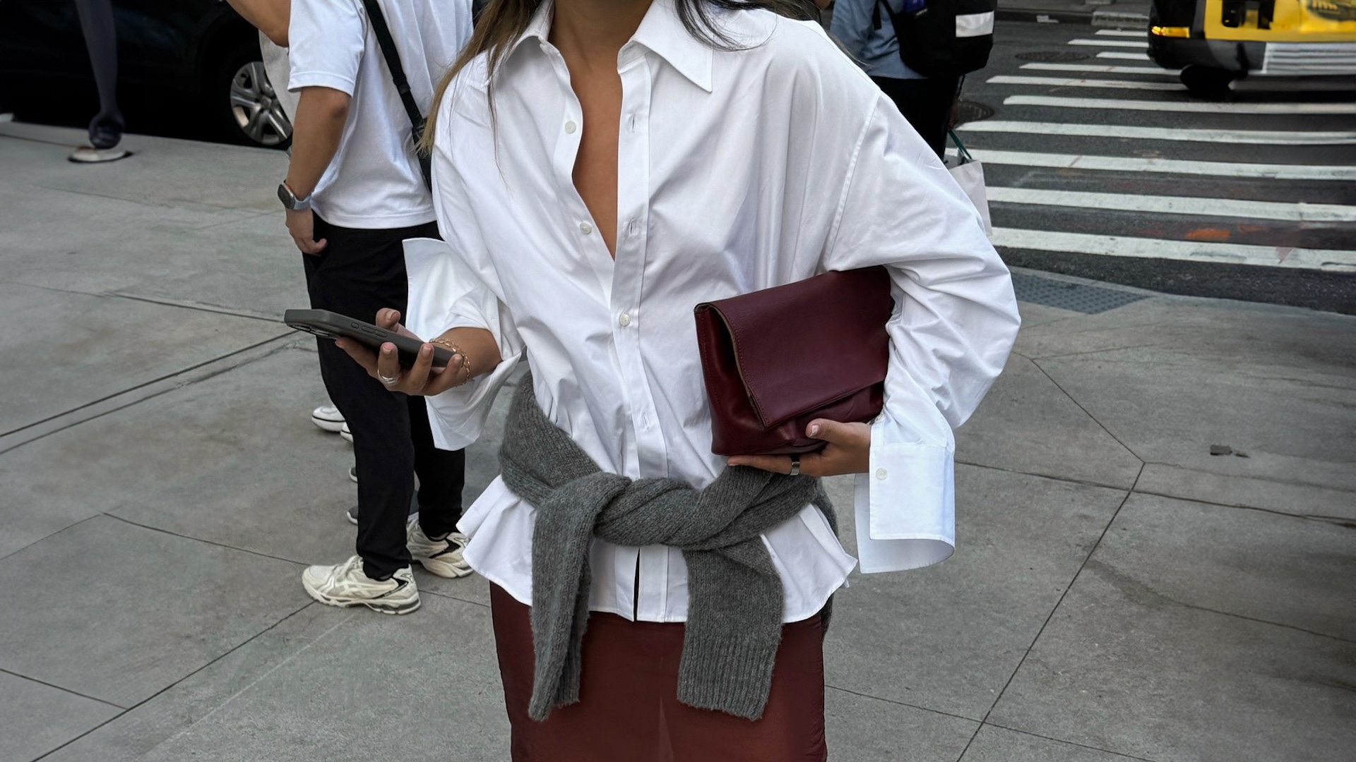 Style influencer Caroline Lin poses in Chicago wearing a white oversize button-down shirt, a burgundy clutch bag, a gray sweater tied around the waist, and a sheer burgundy skirt.
