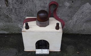 Concrete wall and floor, art piece, white box with small front opening, red cord handle, three dark wood dome pieces on top