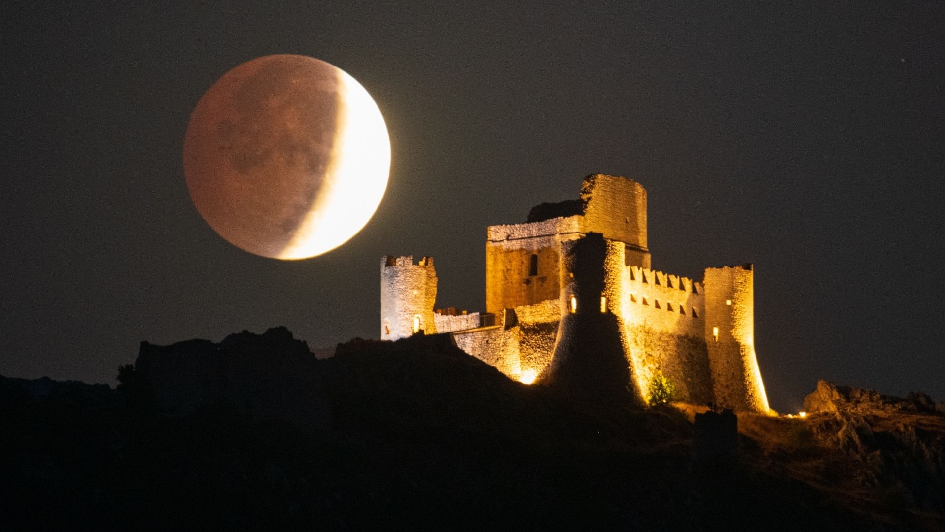A full moon is photographed during a lunar eclipse, as Earth's shadow darkens the lunar disk close to a stone castle on a hill.