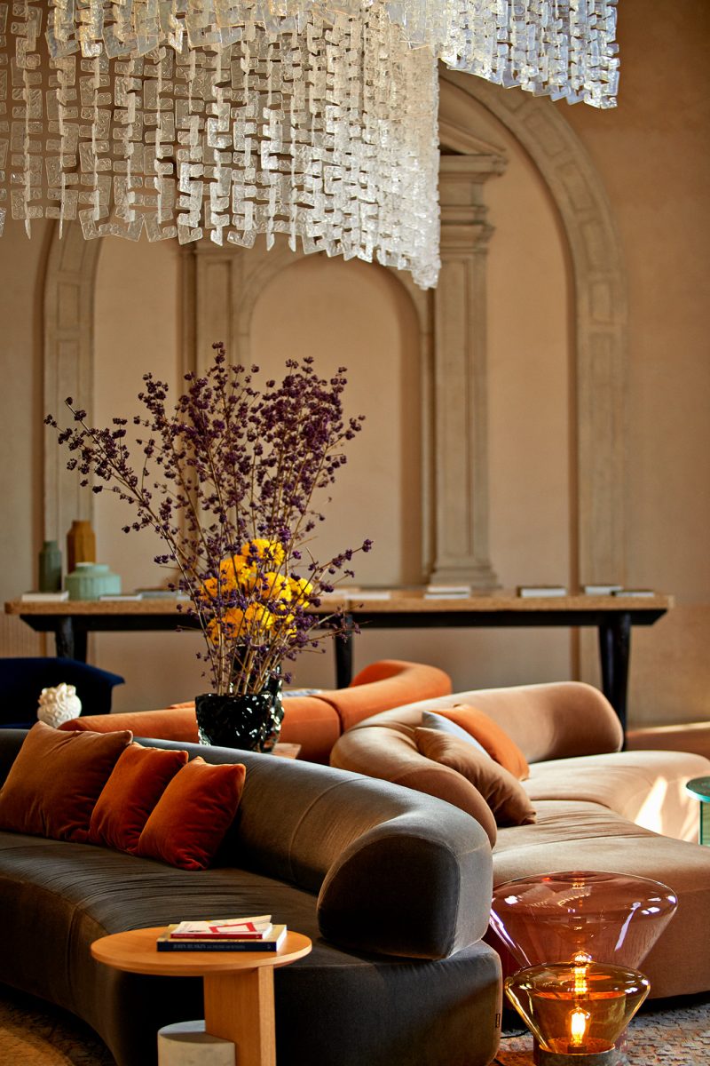 A hotel lobby features wavy furniture designs, including upholstered sofas in earthy tones, round coffee tables, and hand-blown glass lamps, as well as flowers.